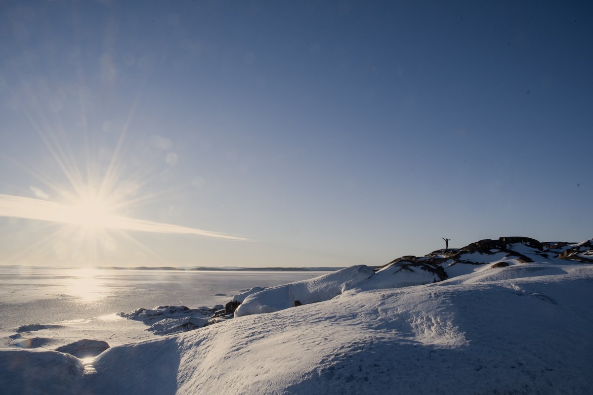 Vy över fruset hav och snötäckta klippor. En människa står högst upp med armarna sträckta utåt och uppåt.
